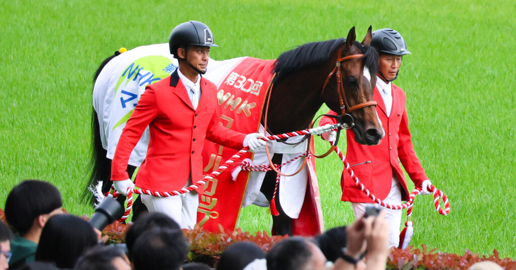 2025年5月11日、東京競馬場で行われたNHKマイルカップの表彰式の様子。JRA職員に引かれたパンジャタワーの首には、優勝レイが掛けられ誇らしげな姿が印象的な一枚