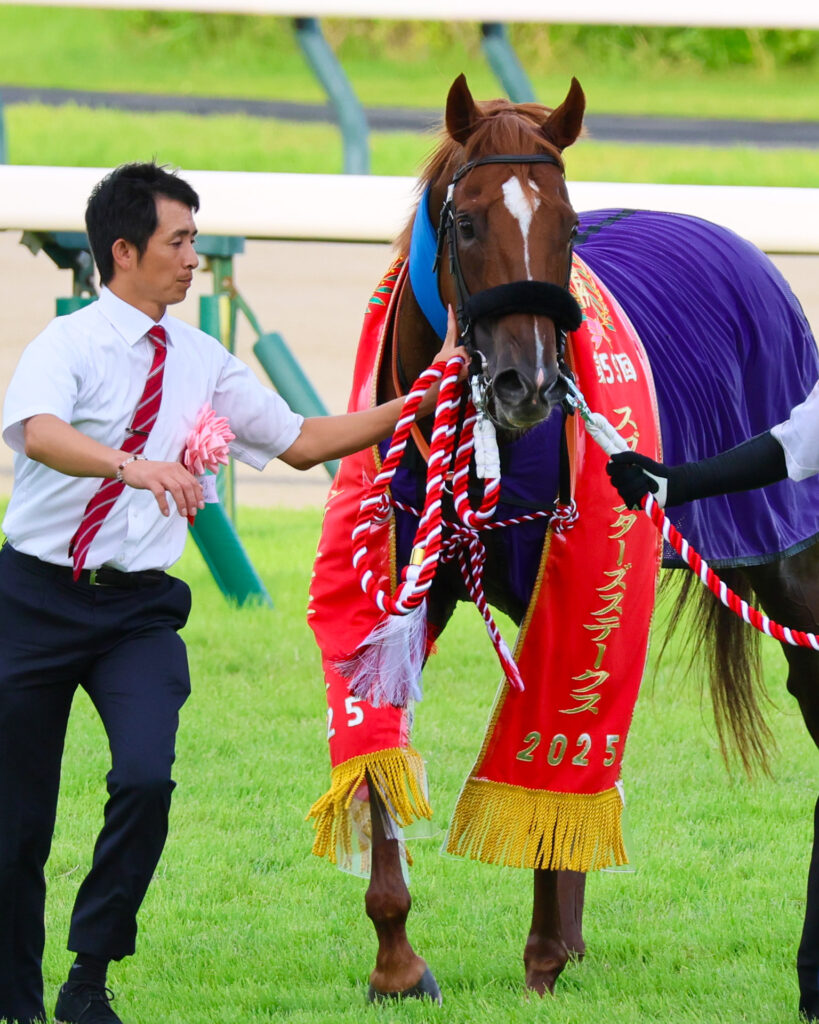 2025年スプリンターズステークス（GⅠ）で勝利を飾ったウインカーネリアンが、中山競馬場で優勝レイを身にまとい立つGⅠ制覇記念の写真。